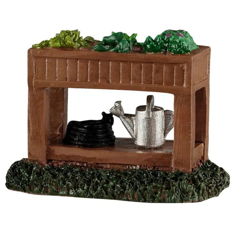 This image shows what this Lemax accessory looks like on a white background. It was designed to look like a small, wooden plant stand. There are plants in the bed up top and on the bottom shelf, there's a black hose and a silver watering can.