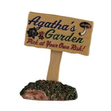This image shows what this Lemax accessory looks like on a white background. It was designed to look like a sign that reads "Agatha's Garden. Pick at your own risk!" There's a mushroom and three flowers on the sign too.
