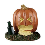 This Lemax accessory looks like a jack-o-lantern on a grassy patch. There's a green bottle on the ground next to the pumpkin. The pumpkin looks like it threw up.