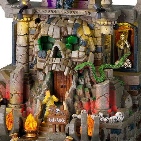 This is an up close view of the lighted building. There's a large green snake hissing at a person trying to enter the cave's doors. There are broken columns near the doors and lots of different stones.
