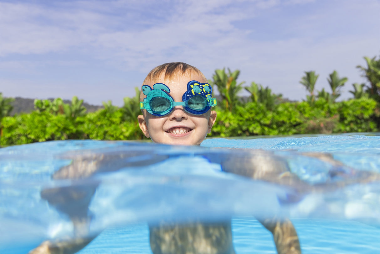 This is a lifestyle image showing a young boy with his head above water, smiling at the camera wearing the turtle themed goggles.
