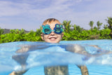 This is a lifestyle image showing a young boy with his head above water, smiling at the camera wearing the turtle themed goggles.