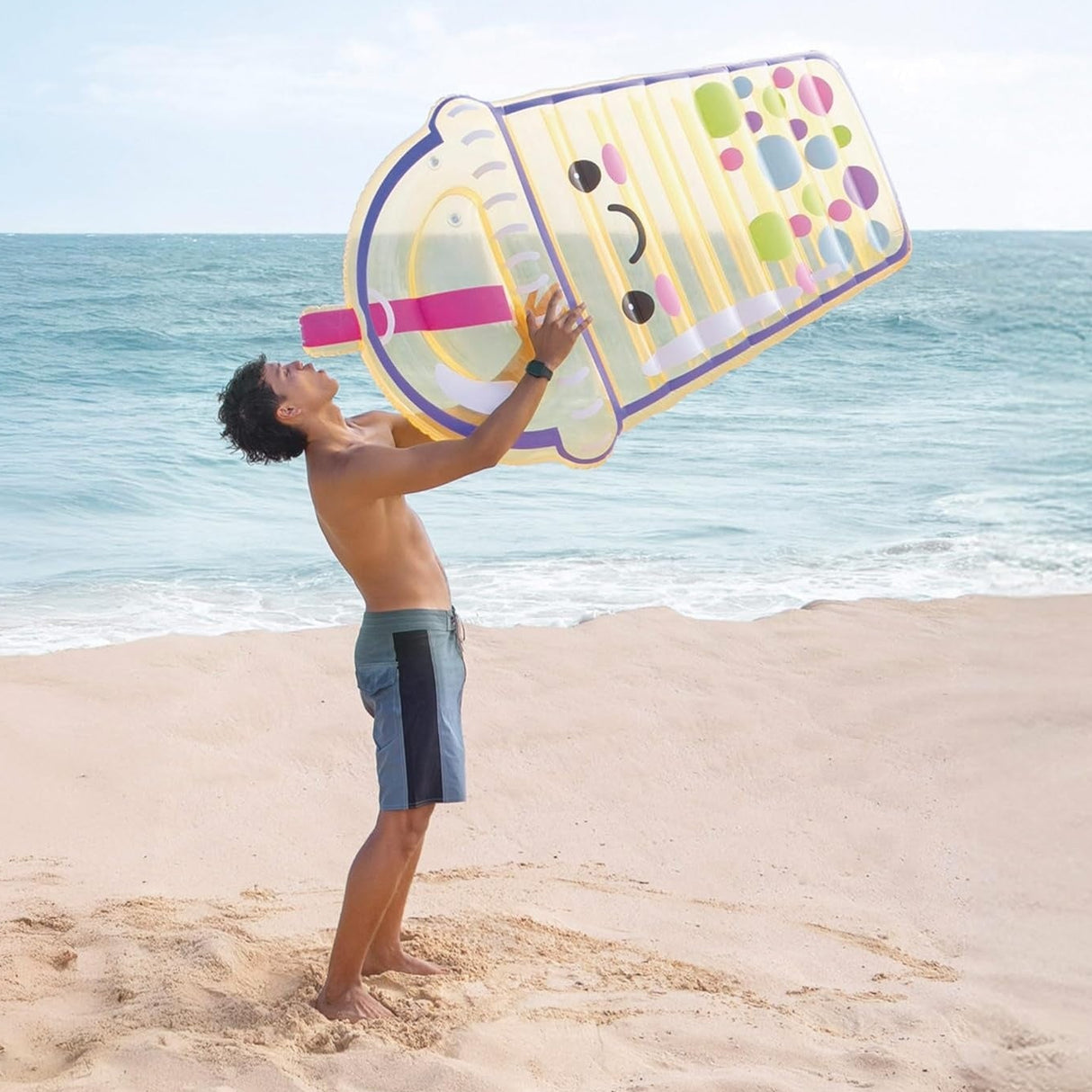 This is a lifestyle image showing a guy standing on the beach holding the boba inflatable pool float like he's taking a sip out of it.