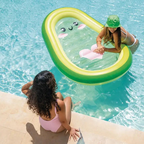 This is a lifestyle image showing a girl leaning on the avocado pool float in a pool while her friend sits on the edge of the pool watching.