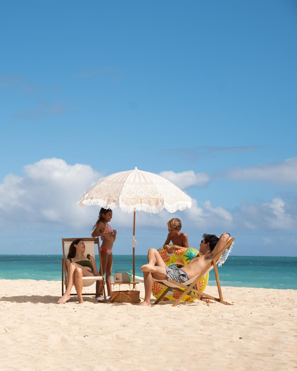 This is a lifestyle image showing a group of people on the beach underneath an umbrella. There's a young boy standing up, leaning against the pineapple inner tube.