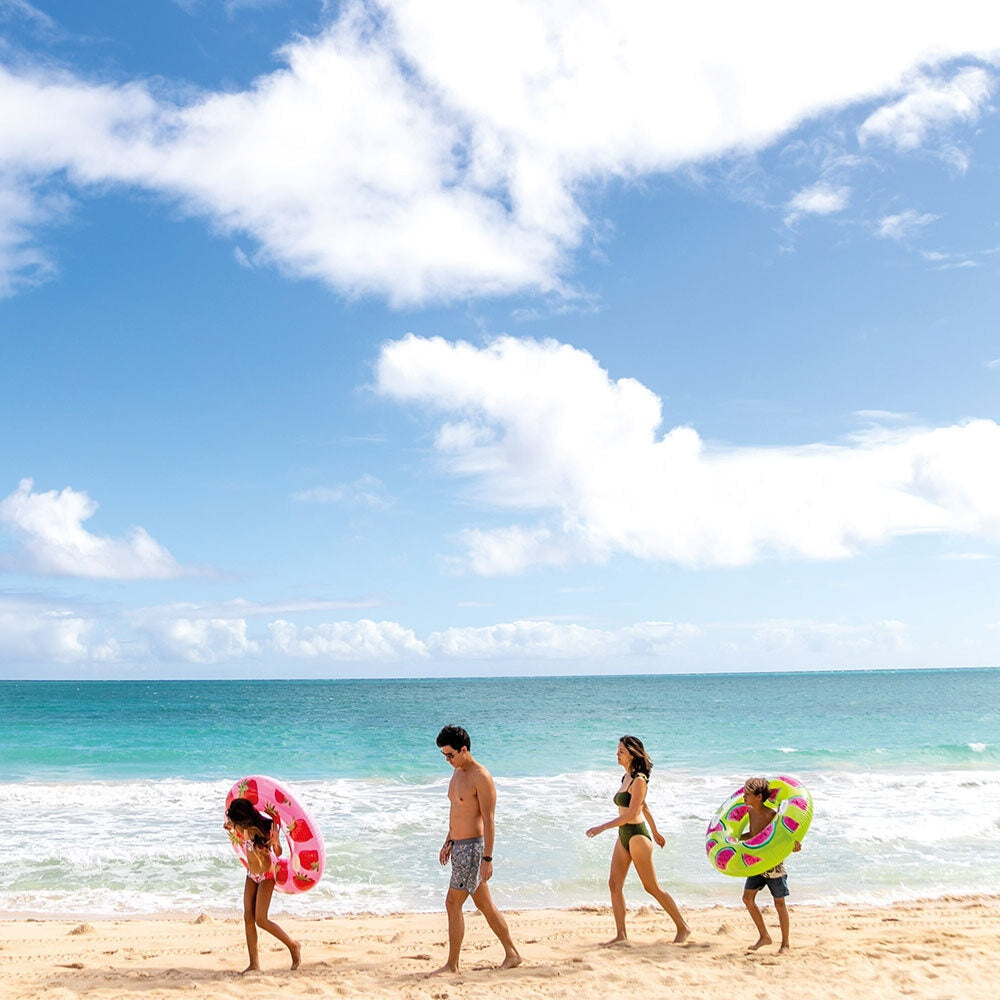 This is a lifestyle image showing people walking along the ocean on a beach. Two of them are holding colorful fruit themed inner tubes.