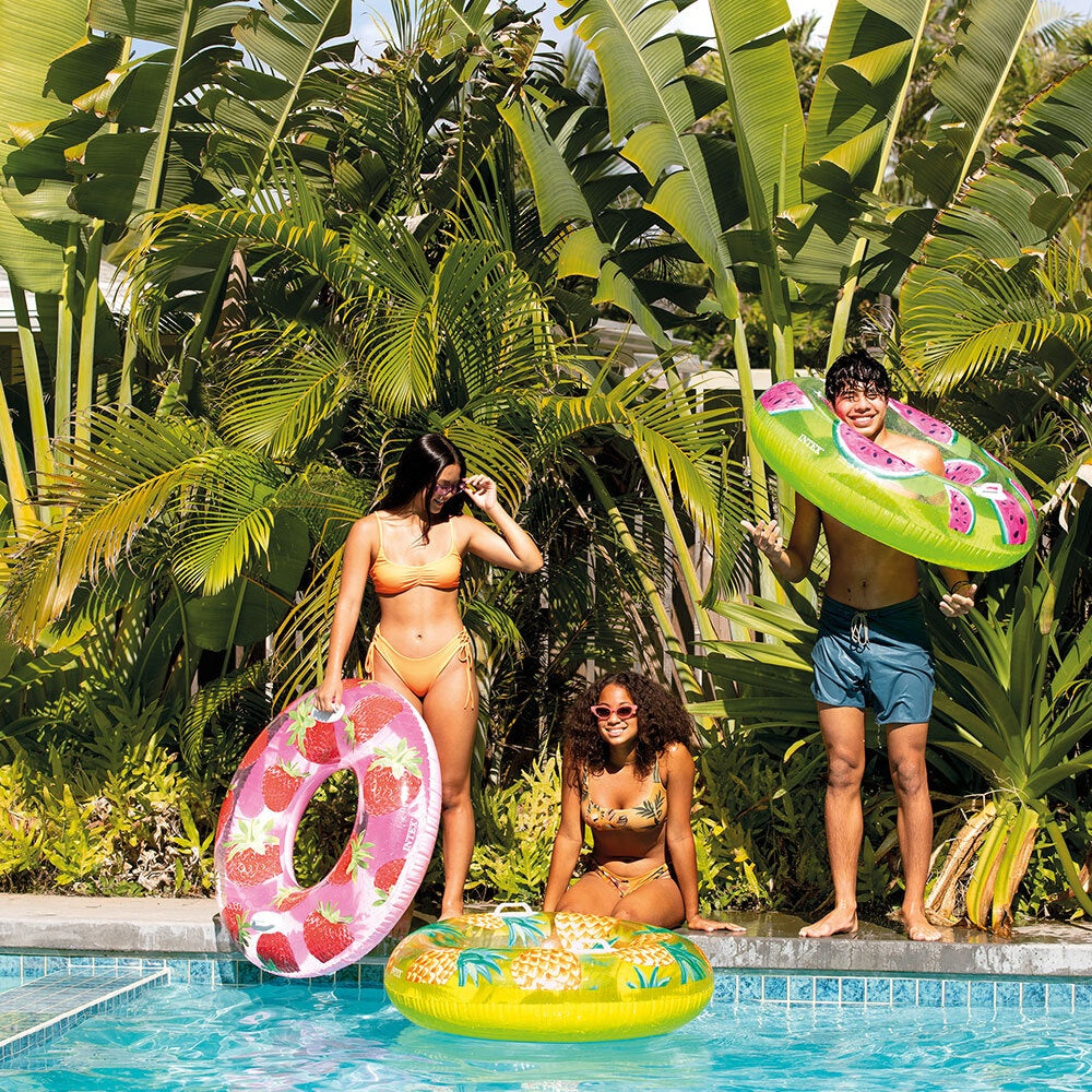 This is a lifestyle image showing two people standing on the edge of a pool while one girl sits. They each have their own fruit themed inner tube.