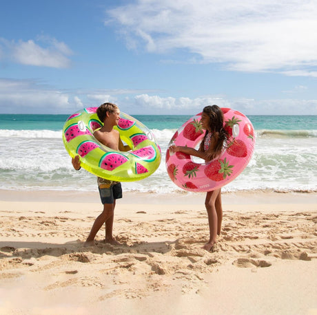 This is a lifestyle image showing two young kids on the beach holding two different inner tubes.