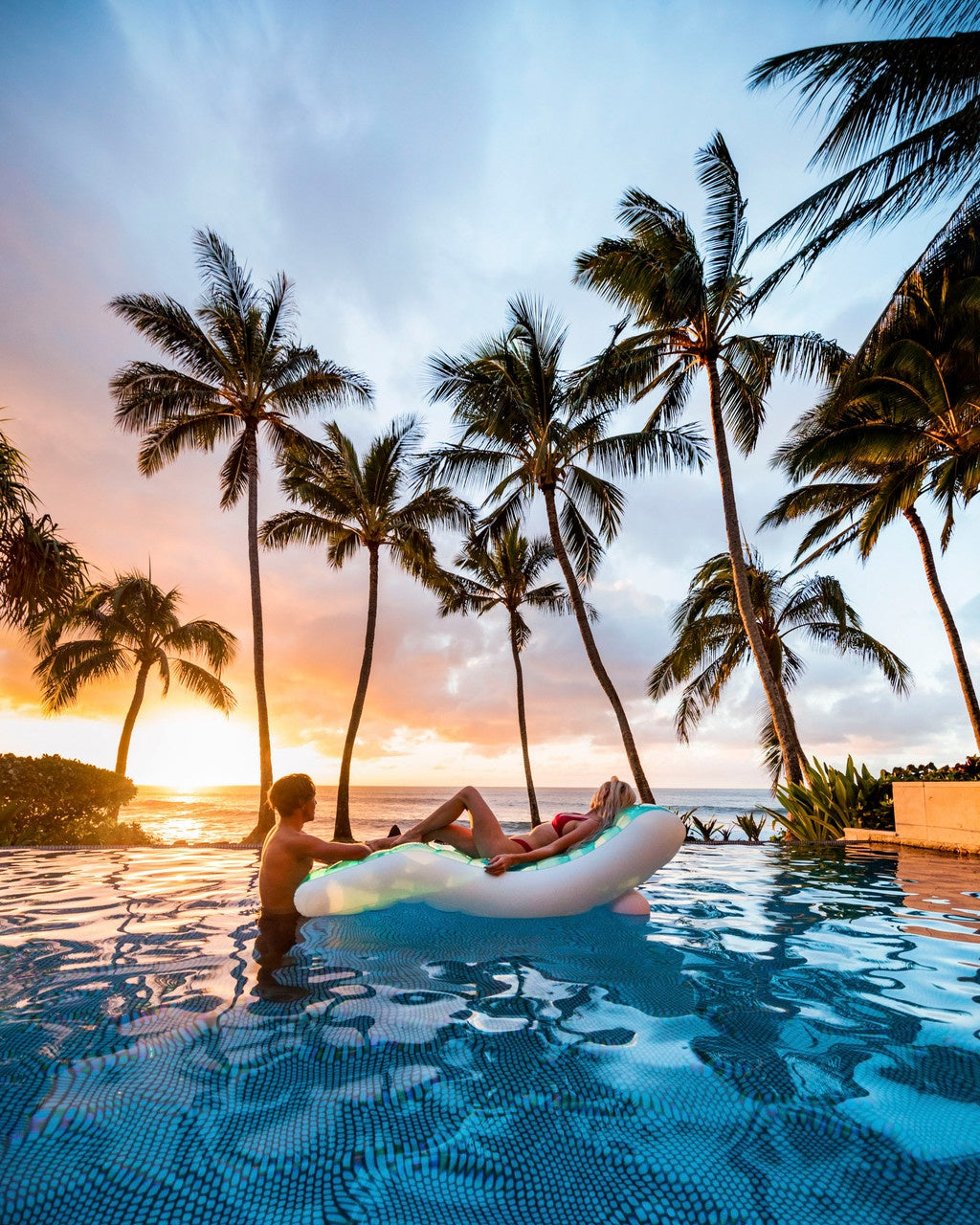 This is a lifestyle image showing a woman sitting on the splash lounge mat in the middle of a pool while a man rests his arm on the bottom of the mat. There's a beautiful sunset and palm trees in the background.