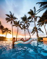 This is a lifestyle image showing a woman sitting on the splash lounge mat in the middle of a pool while a man rests his arm on the bottom of the mat. There's a beautiful sunset and palm trees in the background.