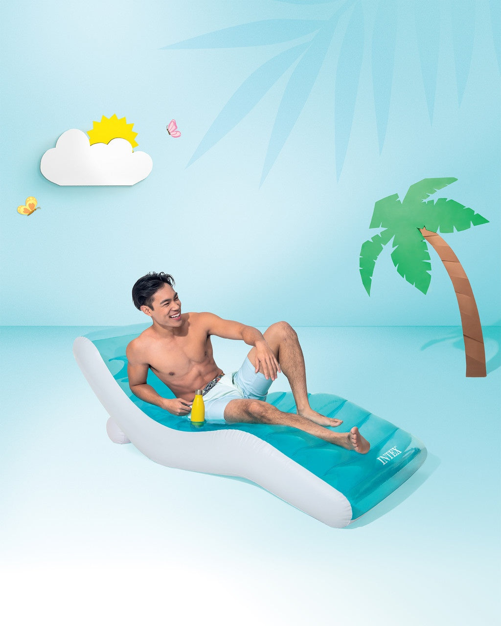 This is a lifestyle image showing a man sitting on the pool chair with a cartoon-ish looking background. The cloud, sun, butterflies and palm tree all look like they're cardboard cutouts.