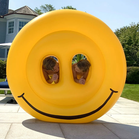 This is a lifestyle image showing the smiley face pool float standing upright on the sidewalk. There's a little boy in the left eye and a little boy in the right eye. They're looking at each other and smiling.