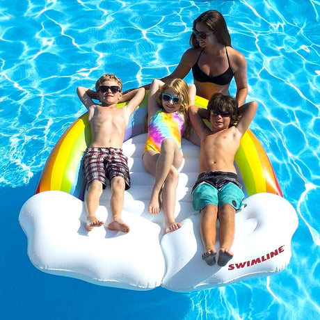 This is a lifestyle image showing three little kids laying on the rainbow pool inflatable while it floats in the pool. A mom is standing behind the float.