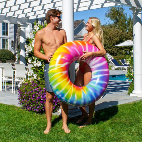 This is a lifestyle image showing a young man and a young woman holding the spiral tie dye swim ring while standing next to a gazebo in a sunny backyard.
