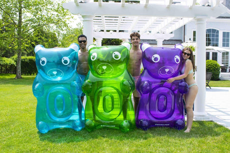 This is a lifestyle image showing three people standing in a backyard underneath a pergola. They're each holding a different colored gummy bear float. From left to right, a young man is holding the blue gummy bear, another young man is holding the green gummy bear and then a young woman is holding the purple gummy bear.