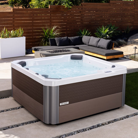 This is a lifestyle image showing this hot tub filled with water on a stone patio next to a fire pit table and an outdoor couch.