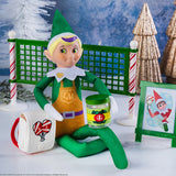 This is a lifestyle image showing a Scout Elf sitting in front of the pickleball net leaning against the backpack and holding the jar of pickles.