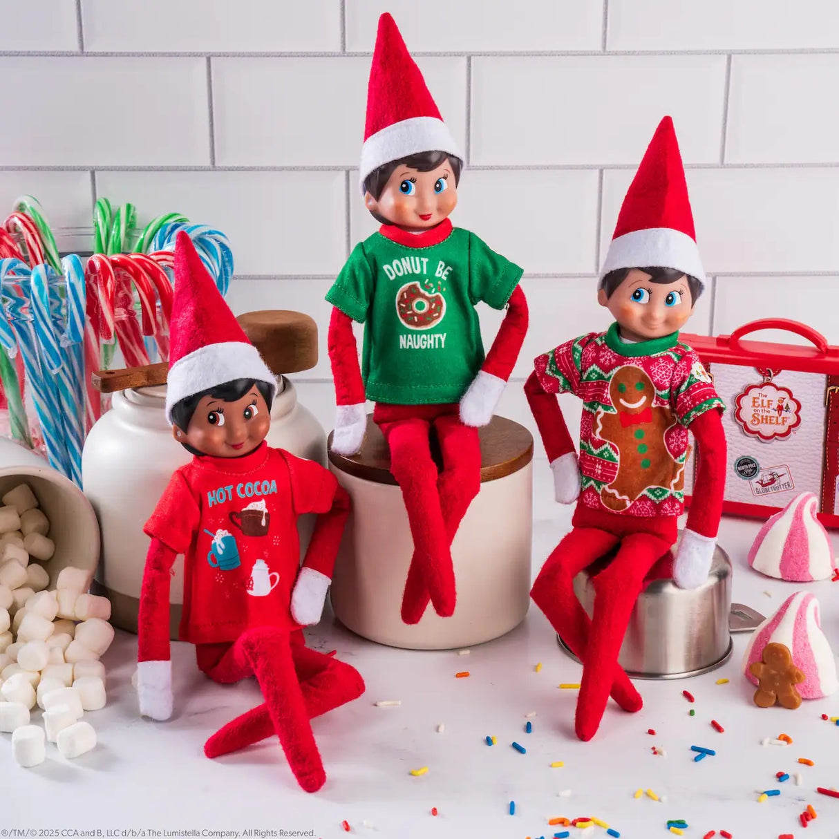 This is a lifestyle image showing three Scout Elves wearing the festive t-shirts. They're set up on a kitchen counter surrounded by multi-colored candy canes, marshmallows, sprinkles, candy and a small gingerbread cookies.