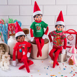 This is a lifestyle image showing three Scout Elves wearing the festive t-shirts. They're set up on a kitchen counter surrounded by multi-colored candy canes, marshmallows, sprinkles, candy and a small gingerbread cookies.