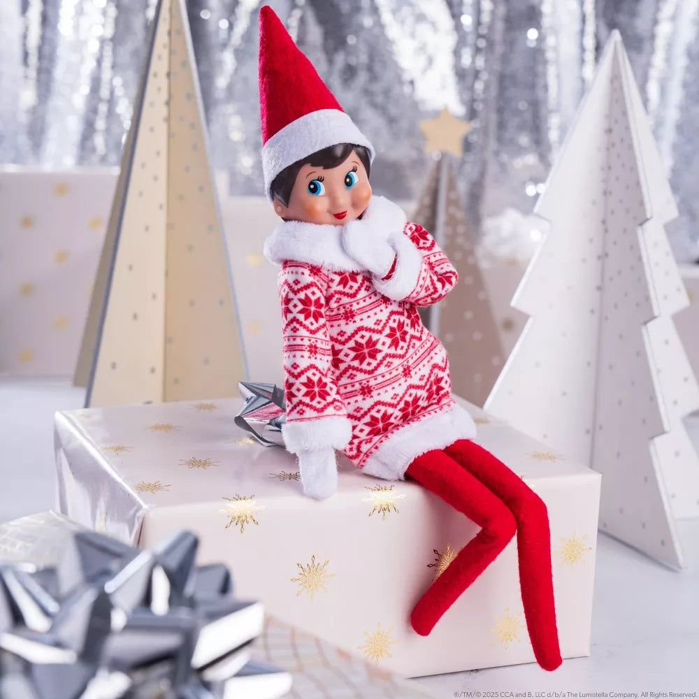 This is a lifestyle image showing a Scout Elf wearing the Sweetie Dress and sitting on a wrapped present.