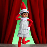 This is a lifestyle image showing a Scout Elf posing in front of red curtains wearing the silver hat accessory and the silver tinsel dress.