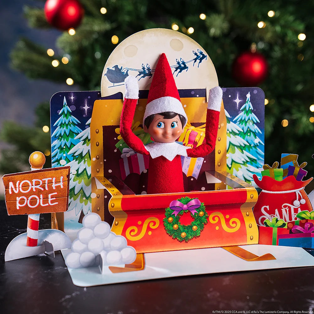 This lifestyle image shows the Sleigh Ride pop up scene. The Scout Elf is in the red sleigh that features gold accents and a green wreath on the front. Behind the sleigh, there are snow covered trees and Santa and his reindeer flying in front of the moon. On the left side, there's a sign that reads "North Pole" and a pile of snowballs. On the right side, there's a big red bag full of Santa's presents.