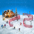 This is a lifestyle image showing this set on display. There's the ski lodge with two trees on either side of it and a sign in front. The fence has been set so skiers can ski in between them and there are lamp posts scattered throughout. There's a couple holding their child's hand as they watch people ski.