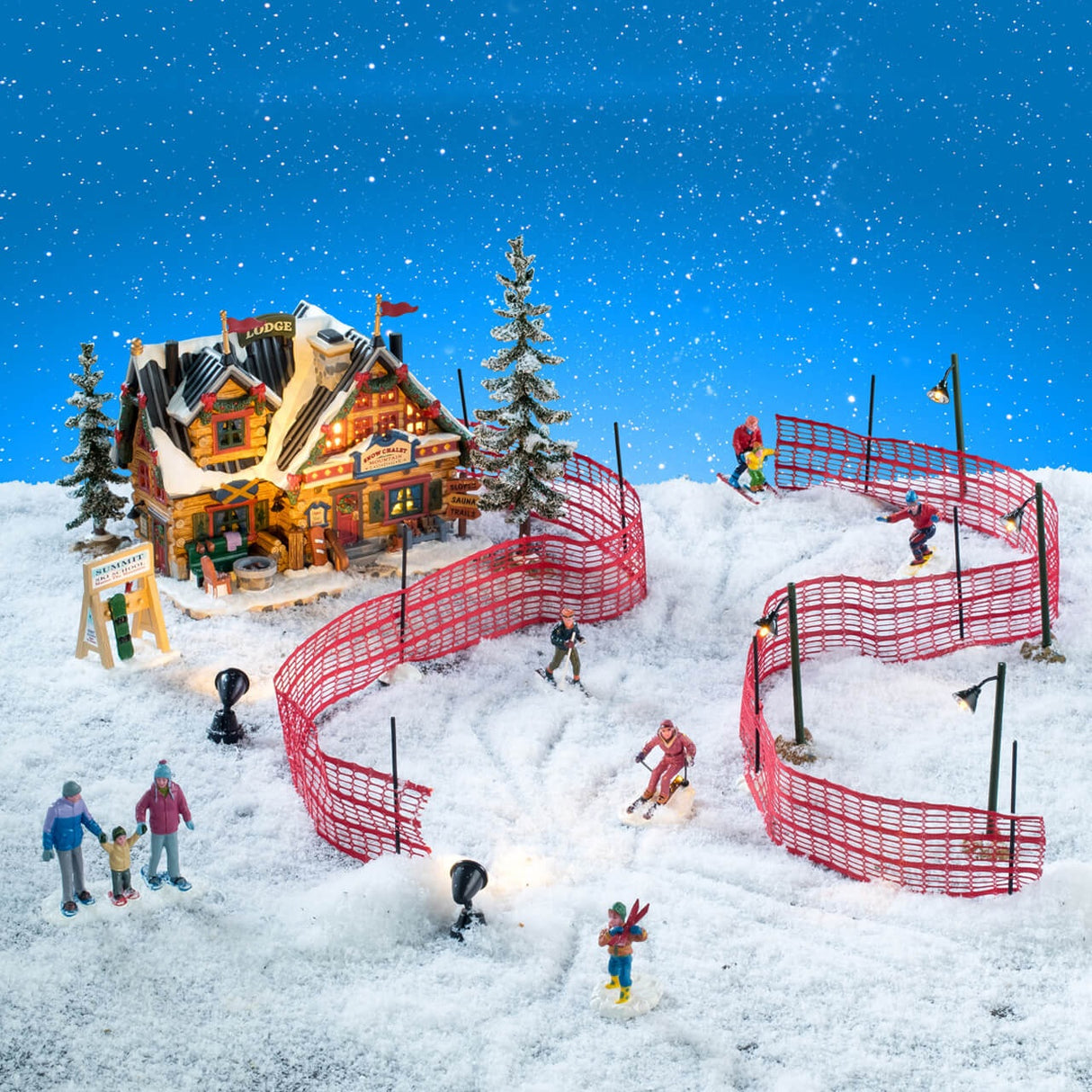 This is a lifestyle image showing this set on display. There's the ski lodge with two trees on either side of it and a sign in front. The fence has been set so skiers can ski in between them and there are lamp posts scattered throughout. There's a couple holding their child's hand as they watch people ski.