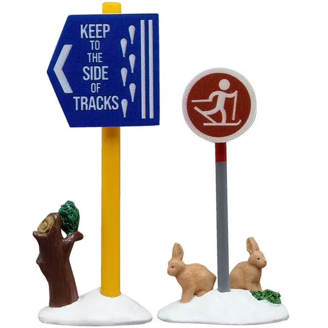 This is a set of two Lemax village signs. One has a yellow pole and the blue sign reads "KEEP TO THE SIDE OF TRACKS" and has a small tree stump next to it. The other sign has a gray pole and a circular, red sign showing a cartoon person skiing and there are two tan colored bunnies underneath it.
