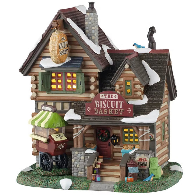 This is a Lemax lighted building designed to look like a tall log cabin. There's a blue bird on the roof and one on the grass near the front stairs. There's snow on the roof and on the grass. There's a sign by the stairs that reads "CREATE YOUR OWN BISCUIT BASKET!" There's a cart on the left of the stairs with biscuits inside. There are jars lining the stairs.
