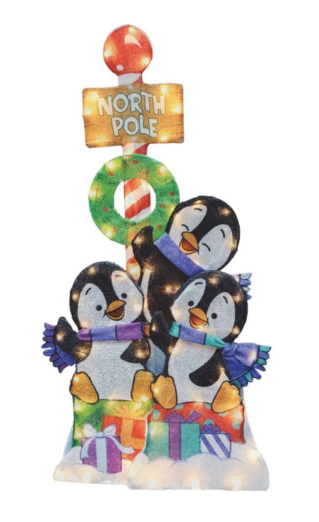 This image shows this decoration on a white background. There are three playful penguins sitting on a bunch of presents. There's a North Pole sign with a wreath behind them. The clear lights are visible.