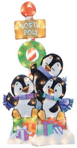 This image shows this decoration on a white background. There are three playful penguins sitting on a bunch of presents. There's a North Pole sign with a wreath behind them.