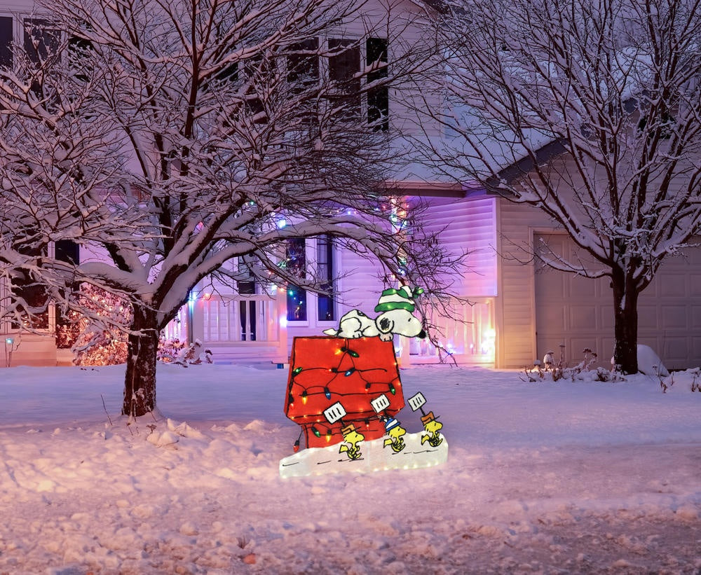 This is a lifestyle image showing the Snoopy doghouse decoration on a snowy front lawn decorated for Christmas.