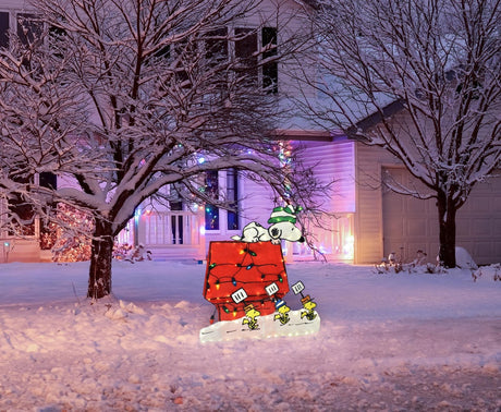 This is a lifestyle image showing the Snoopy doghouse decoration on a snowy front lawn decorated for Christmas.