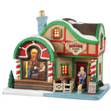 This is a Lemax lighted building designed to look like a small cottage turned into a holiday themed cafe. The roof and sidewalk are covered in snow. There's a fenced in area with a reindeer. A woman is leaving the cafe with a dog on a leash and the dog is wearing antlers. There's a big window in the front you can see people ordering from the register. There are red and white striped columns and green accents.