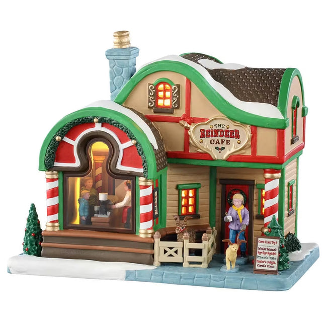 This is a Lemax lighted building designed to look like a small cottage turned into a holiday themed cafe. The roof and sidewalk are covered in snow. There's a fenced in area with a reindeer. A woman is leaving the cafe with a dog on a leash and the dog is wearing antlers. There's a big window in the front you can see people ordering from the register. There are red and white striped columns and green accents.