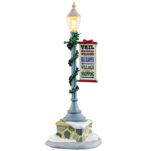 This is a Lemax village accessory designed to look like a lamp post. It's on a stone base covered in snow. The post is wrapped with shiny green garland. There's a sign hanging off the post that reads "VAIL Holiday WELCOME. SKI SLOPES (to the left). VILLAGE and SHOPPING (to the right)."