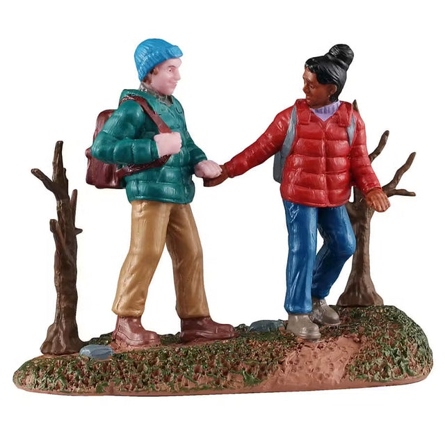 This is a Lemax figurine designed to look like a man and a woman holding hands on a hike. He's wearing a bright blue winter hat, a teal coat, brown pants and brown shoes and is wearing a brown backpack. She has her black hair in a bun and is wearing a red coat over a purple shirt, blue jeans and brown shoes. She is wearing a gray backpack. They're walking on grass in between two small, bare trees.
