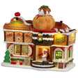 This is a Lemax lighted building designed to look like it was made out of gingerbread, waffles, cookies, frosting and sprinkles. There are signs on the building that read "TIME TO EAT!" and "BAKED FRESH!" There are signs on the sidewalk that read "SPECIAL: Apple Dumpling a la Mode with Salted Caramel Sauce" and "THE ORCHARD TIMES." There's a gingerbread man standing near the front door holding a plate. Inside the window, you can see a gingerbread person waiting for their food.