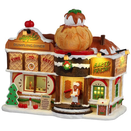 This is a Lemax lighted building designed to look like it was made out of gingerbread, waffles, cookies, frosting and sprinkles. There are signs on the building that read "TIME TO EAT!" and "BAKED FRESH!" There are signs on the sidewalk that read "SPECIAL: Apple Dumpling a la Mode with Salted Caramel Sauce" and "THE ORCHARD TIMES." There's a gingerbread man standing near the front door holding a plate. Inside the window, you can see a gingerbread person waiting for their food.