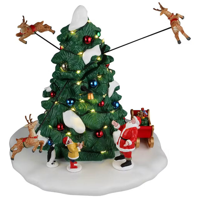 This is a Lemax table accent designed to look like a tall, decorated Christmas tree. There are three reindeer flying around it. Santa, a young boy and a penguin stand at the base of the tree. There's also a red and gold sleigh with presents inside. There's snow on the ground and on the tree.