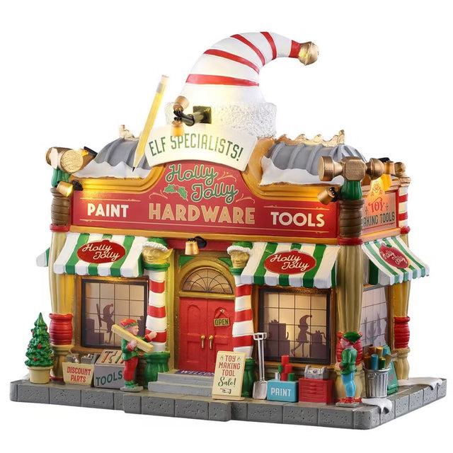 This is a Lemax lighted building designed to look like a hardware store with a giant elf hat statue on the roof. There's snow on the roof and on the sidewalk. Along the front of the store, there's a potted tree, signs that read "DISCOUNT PARTS," "TOOLS," "TOY MAKING TOOL SALE!" and "PAINT." Inside, you can see elves working.