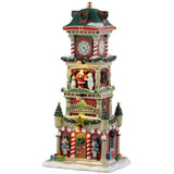 This is a Lemax lighted building designed to look like a clock tower decorated for Christmas. There are garlands and wreaths and decorated Christmas trees everywhere. On the second floor of the tower, there's a snowman, Santa and a polar bear standing on the balcony. On either side of the door, there are red and white striped columns and nutcrackers. There's snow on the roof and on the sidewalk.