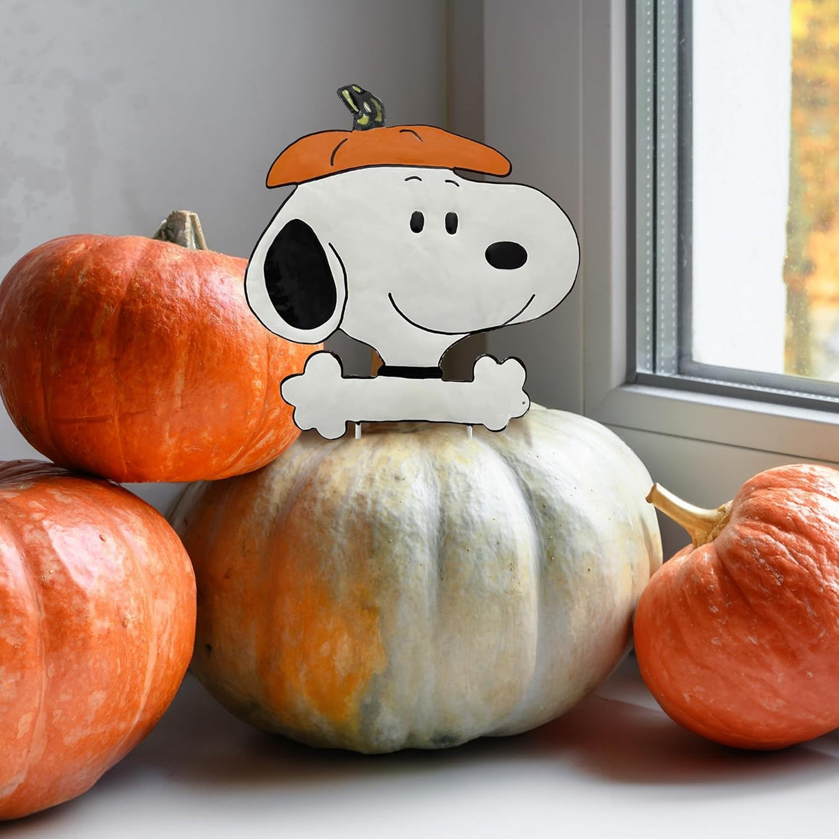 This is a lifestyle image showing off this pumpkin press-in decoration. It's sticking out of a white colored pumpkin near a windowsill. The pumpkin press-in decoration is designed to look like the famous black and white beagle from Peanuts cartoons: Snoopy. He's wearing the top of a pumpkin as a hat.