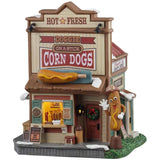 This is a Lemax lighted building designed to look like a tall shack. At the top of the building, it reads "HOT & FRESH. DOGGIE ON A STICK CORN DOGS." There's a large sideways corndog statue on the roof. There's a booth you can walk up to and grab some corn dogs and condiments. There's a wreath on the front door and a large corndog statue wearing a hat and gloves. There's a goofy face on the corndog statue and he's waving.