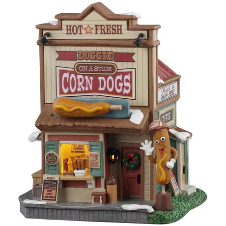This is a Lemax lighted building designed to look like a tall shack. At the top of the building, it reads "HOT & FRESH. DOGGIE ON A STICK CORN DOGS." There's a large sideways corndog statue on the roof. There's a booth you can walk up to and grab some corn dogs and condiments. There's a wreath on the front door and a large corndog statue wearing a hat and gloves. There's a goofy face on the corndog statue and he's waving.
