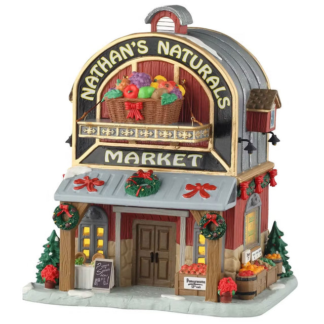 This is a Lemax lighted building designed to look like a red and gray colored barn. The sign up top reads "NATHAN'S NATURALS MARKET" and has a large statue of a basket of fruit on it. There are decorated trees and wreaths. Near the front door, there's a table with chairs and crates and barrels of fruits along the sidewalk.