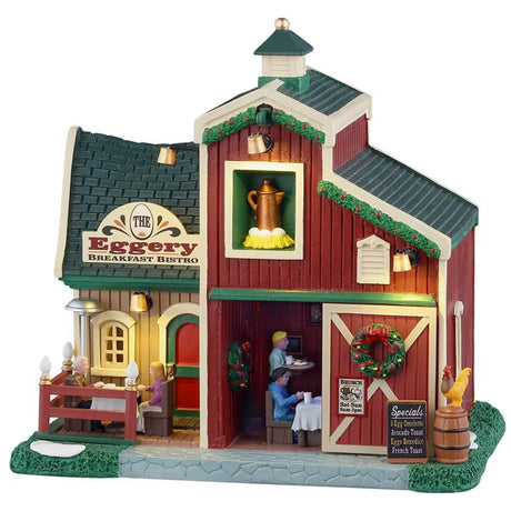 This is a Lemax lighted building designed to look like a barn turned into a breakfast restaurant. There are decorated garlands along the roof and a decorated wreath on the sliding barn door. There's a couple eating at a table on the balcony. Inside the building, there's a waiter bringing a table of people their breakfast. There's some snow on the grass.
