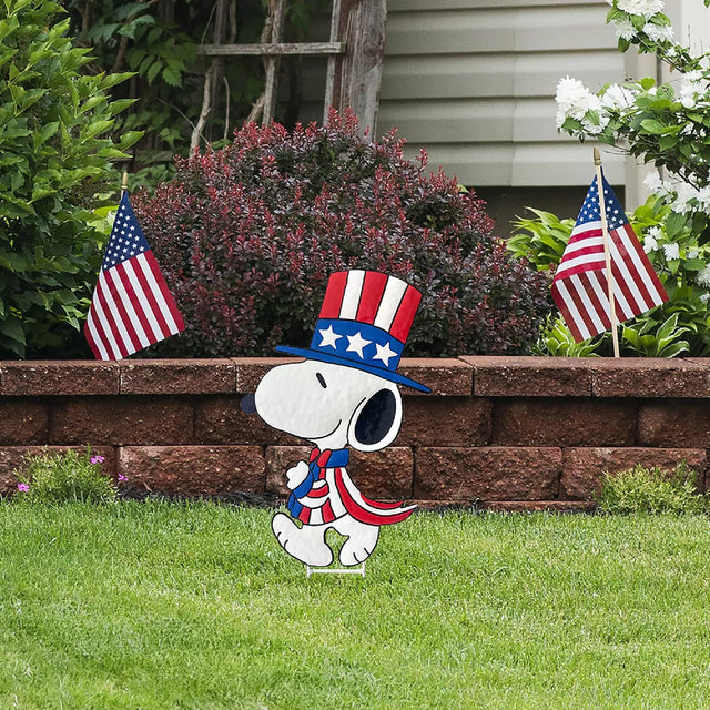 This lifestyle image shows this decorative garden stake shaped like the famous Peanuts character, Snoopy. He's wearing a red, white and blue top hat and suit.