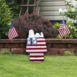 This is a lifestyle image showing this decorative garden stake in a garden. This decorative garden stake is designed to look like the famous Peanuts character, Snoopy. Snoopy is sleeping on top of his doghouse and the house is designed to look like the American flag: red, white and blue stars and stripes.